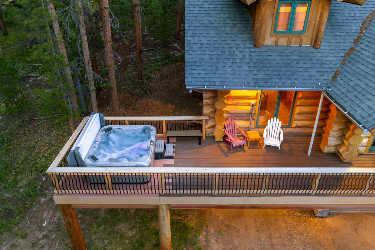 A spacious wooden deck extends from a rustic log cabin, complete with a hot tub, comfortable chairs, and panoramic views of the surrounding forest. The area offers ample space for socializing and soaking in the scenery, while the warm wood tones create a welcoming vibe.