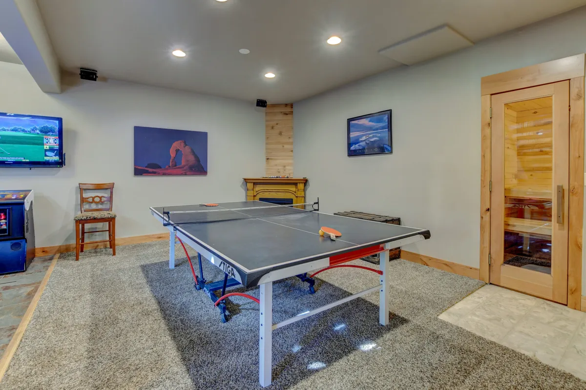 This image captures a lively recreation space with a ping pong table at the center, a wall-mounted television, visible sauna, and eye-catching artwork. The arrangement suggests a versatile area ideal for both active fun and relaxation, reflecting the spirit of multi-generational entertainment.
