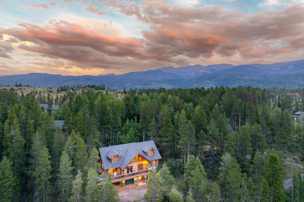 A log cabin-style home is nestled against a backdrop of dense forest and dramatic mountain peaks under a colorful sunset sky. The inviting setting hints at hiking, nature walks, and outdoor gatherings that guests can enjoy right from their private retreat.