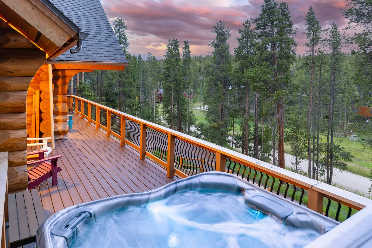 A beautifully designed wooden deck surrounded by lush trees features a bubbling hot tub as its centerpiece. Comfortable outdoor furniture is arranged to maximize relaxation, and the scene is framed by an open sky with some clouds. The tranquil atmosphere hints at both privacy and connection with nature.