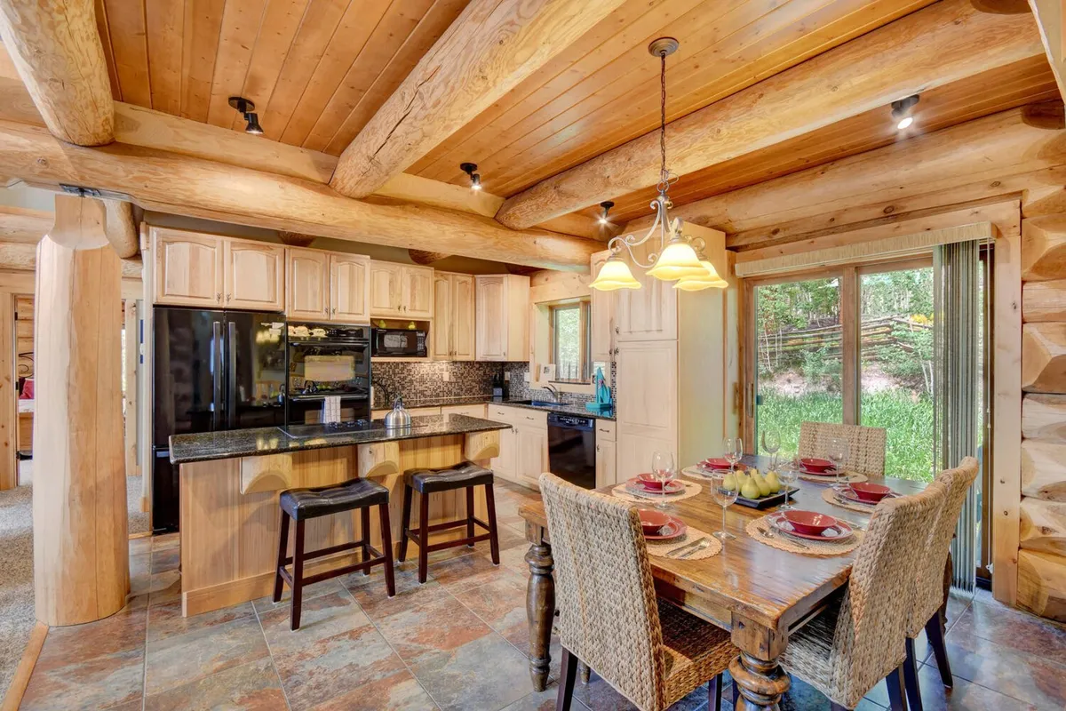 A welcoming dining table is set for a meal, adjacent to a kitchen island with bar stools. Large windows let in natural light and offer glimpses of the outdoors, reinforcing the connection between indoor comfort and the mountain landscape beyond.