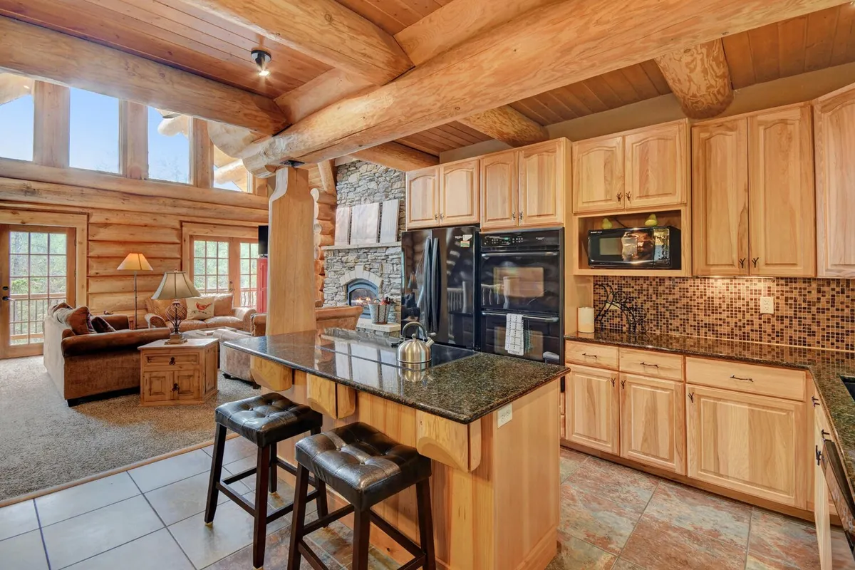 The open-concept living space blends the kitchen, dining, and living areas. It showcases a central kitchen island, log beams overhead, a stone fireplace, and spacious living area seating. Expansive windows bring in mountain views, further enhancing the sense of openness and connection.