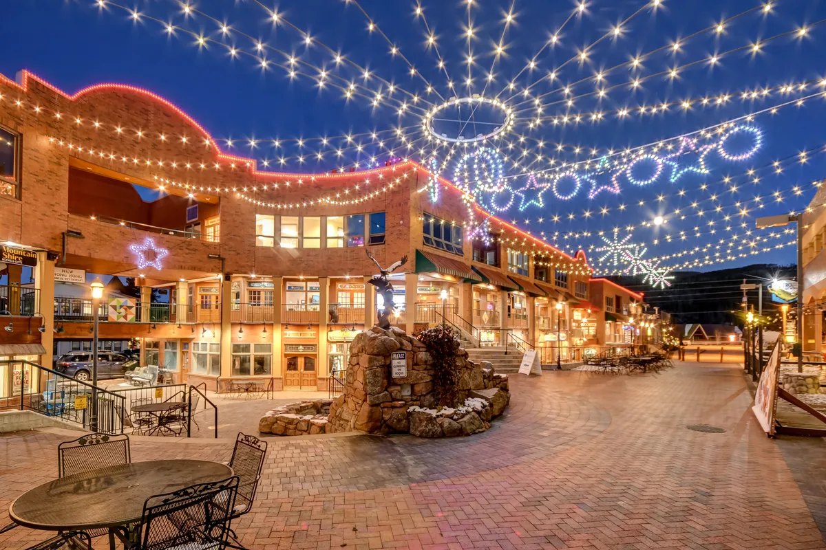 A welcoming streetscape at dusk, adorned with string lights and decorative stars, leading to an outdoor seating area beside a fountain. The scene is alive with people enjoying evening strolls, dining al fresco, and shopping, evoking a joyful, inviting small-town atmosphere.