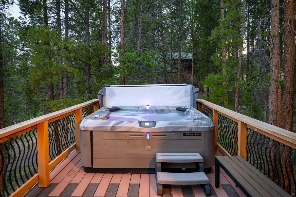 A serene outdoor hot tub set on a wooden deck, surrounded by tall trees. The tub is steaming, inviting guests to relax and enjoy the fresh mountain air. The secluded setting emphasizes connection with nature and opportunities for stargazing or wildlife watching.