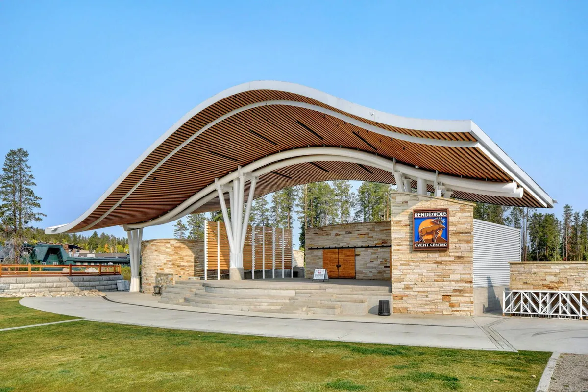 A lively outdoor stage set against a modern architectural backdrop, featuring a curved roof and stone façade. The area is abuzz with people gathered for a community event or concert, capturing the festive spirit of local celebrations and seasonal festivals.