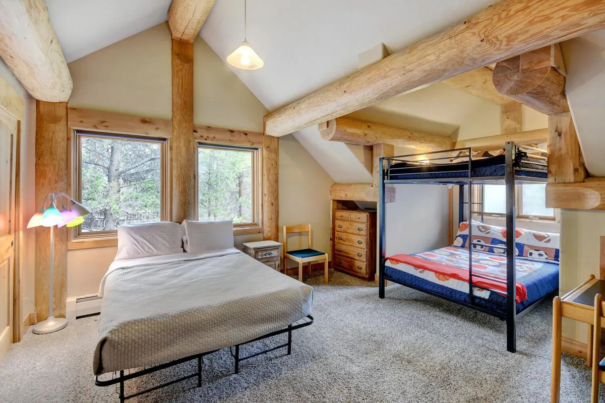 A spacious family-friendly bedroom with a main bed dressed in white linens, a colorful bunk bed, large windows filling the room with light, and exposed wooden beams and logs, creating a warm and welcoming environment.