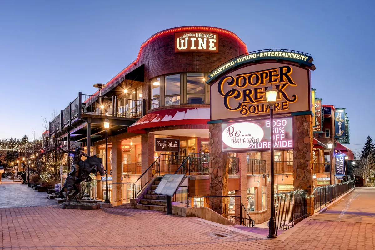 A bustling outdoor square featuring the Cooper Creek Square sign, a wine shop, outdoor seating, and a prominent horse sculpture. The lively setting suggests ongoing dining, shopping, socializing, and entertainment, with hints of seasonal festivals and community activities.