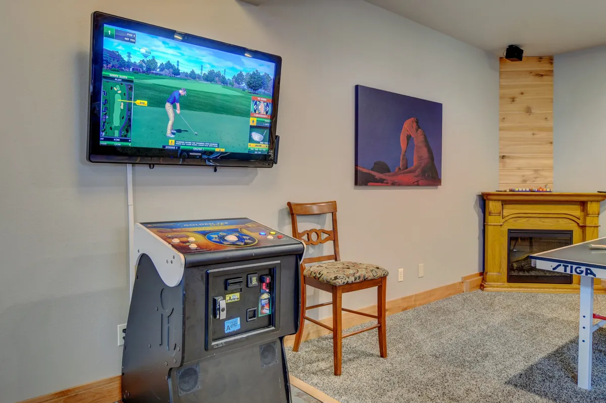 The focus here is on the entertainment zone, featuring an arcade game machine, a television with a golf video game displayed, a wooden chair, a cozy fireplace, and decorative touches. The mix of digital and classic gaming elements, along with the inviting fire, conveys comfort and nostalgic fun.