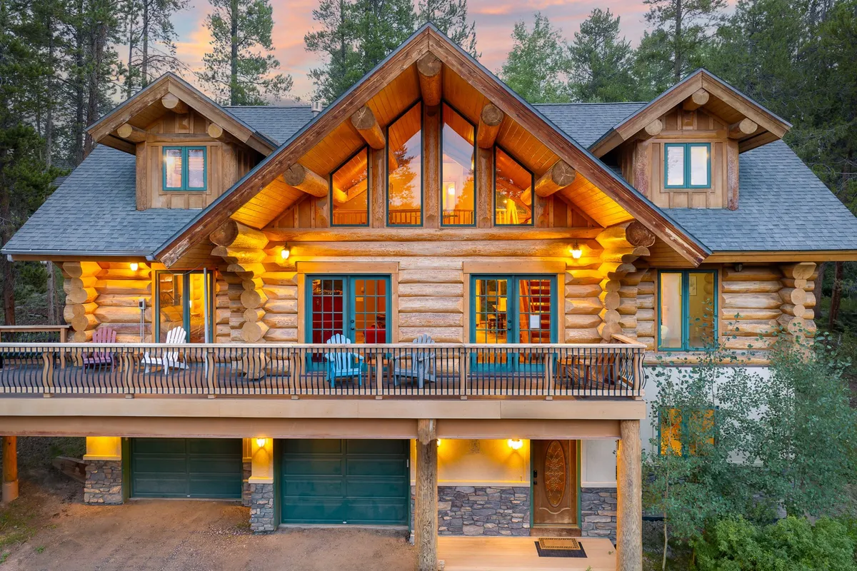 A cozy log cabin exterior featuring a spacious deck with comfortable seating, large windows, and stone accents. The scene suggests an inviting spot for group gatherings, family conversations, or simply soaking in the mountain atmosphere.