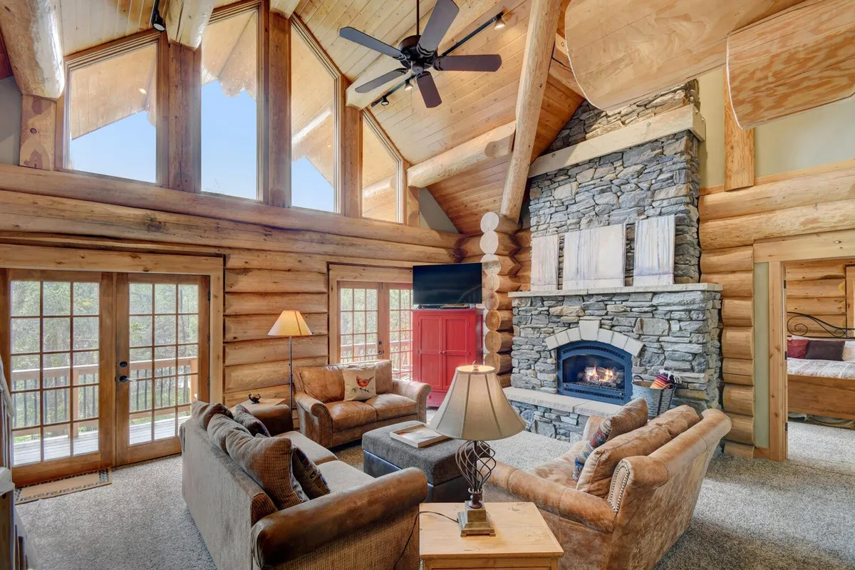 This image features a stunning great room anchored by a grand stone fireplace, surrounded by comfortable seating. Exposed wooden beams and a decorative lamp underscore the rustic yet elegant design, while large windows flood the space with natural light and offer scenic views.