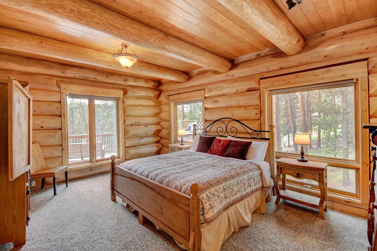 A rustic bedroom featuring a wooden bed, large windows letting in natural light, and log walls. The nightstand with a lamp adds a cozy touch, and the bedding appears plush and inviting.